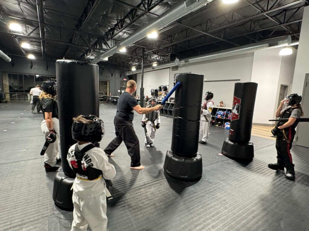 Martial arts instructor demonstrating striking techniques while students practice on training equipment in a beginner class environment, learning martial arts class etiquette for beginners